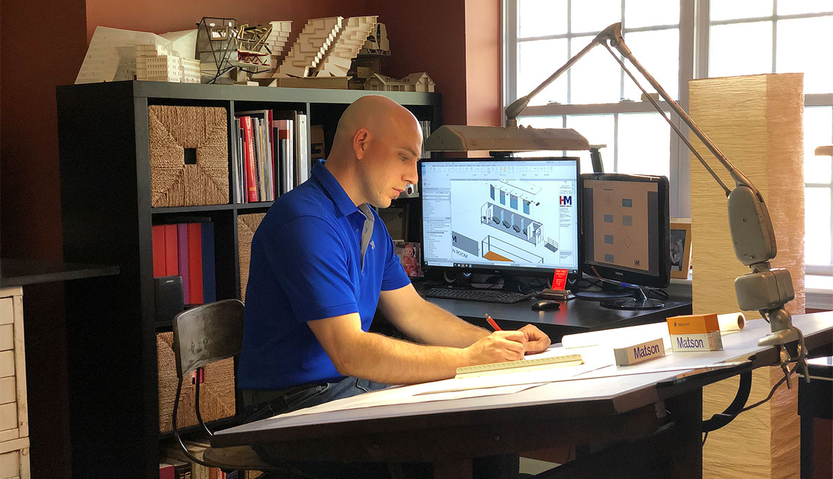 Architect alumnus Erik Fred at work at his drafting desk with a monitor over his shoulder displaying an architectural rendering 