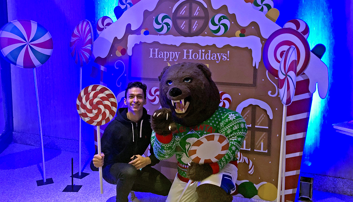 Student Malik Asad with the NYIT Bear in front of holiday decor. 