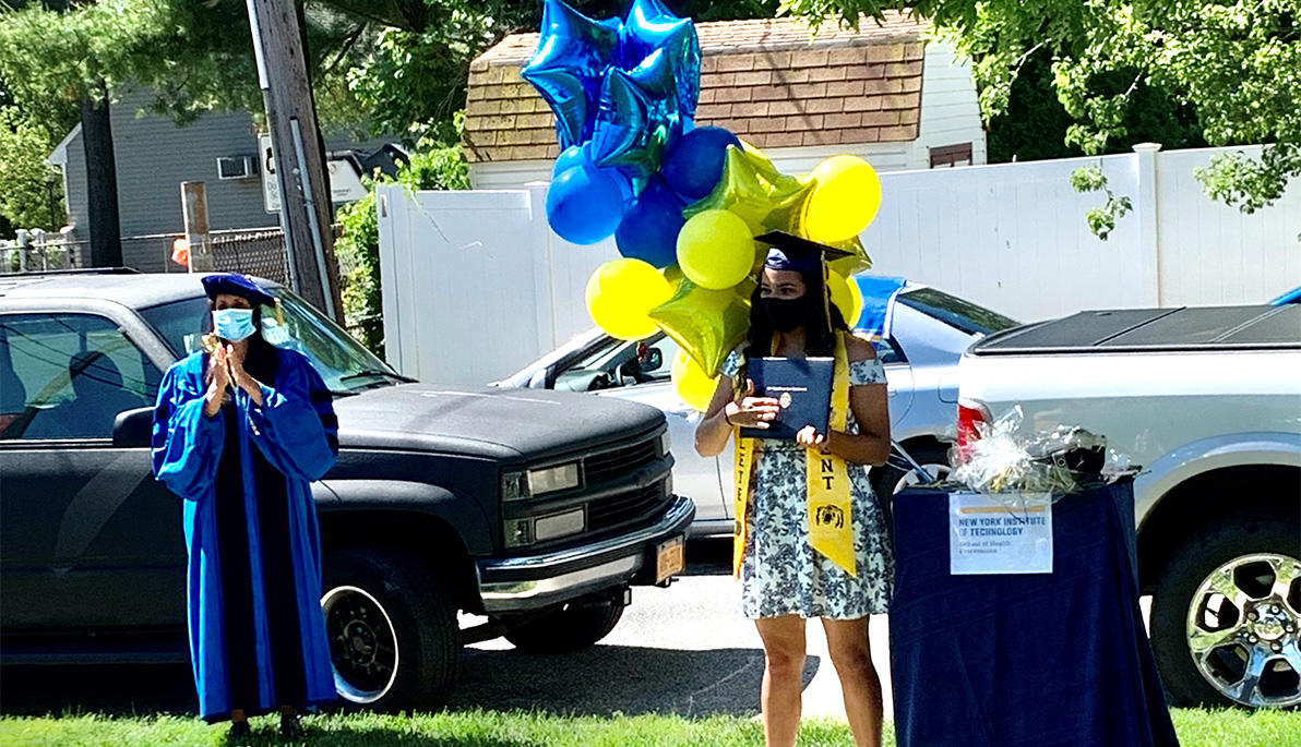 Masked student in mortarboard and surrounded by balloons receives her diploma.