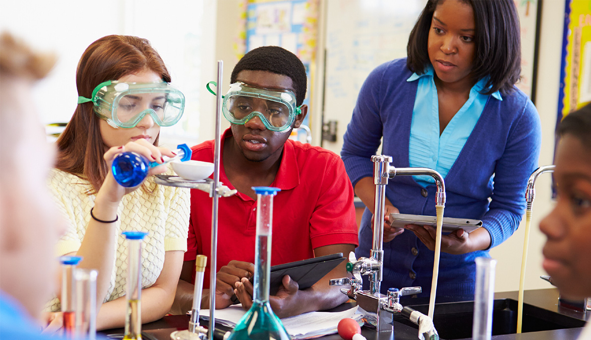 Students in goggles engaging in lab experiment under the supervision of a teacher