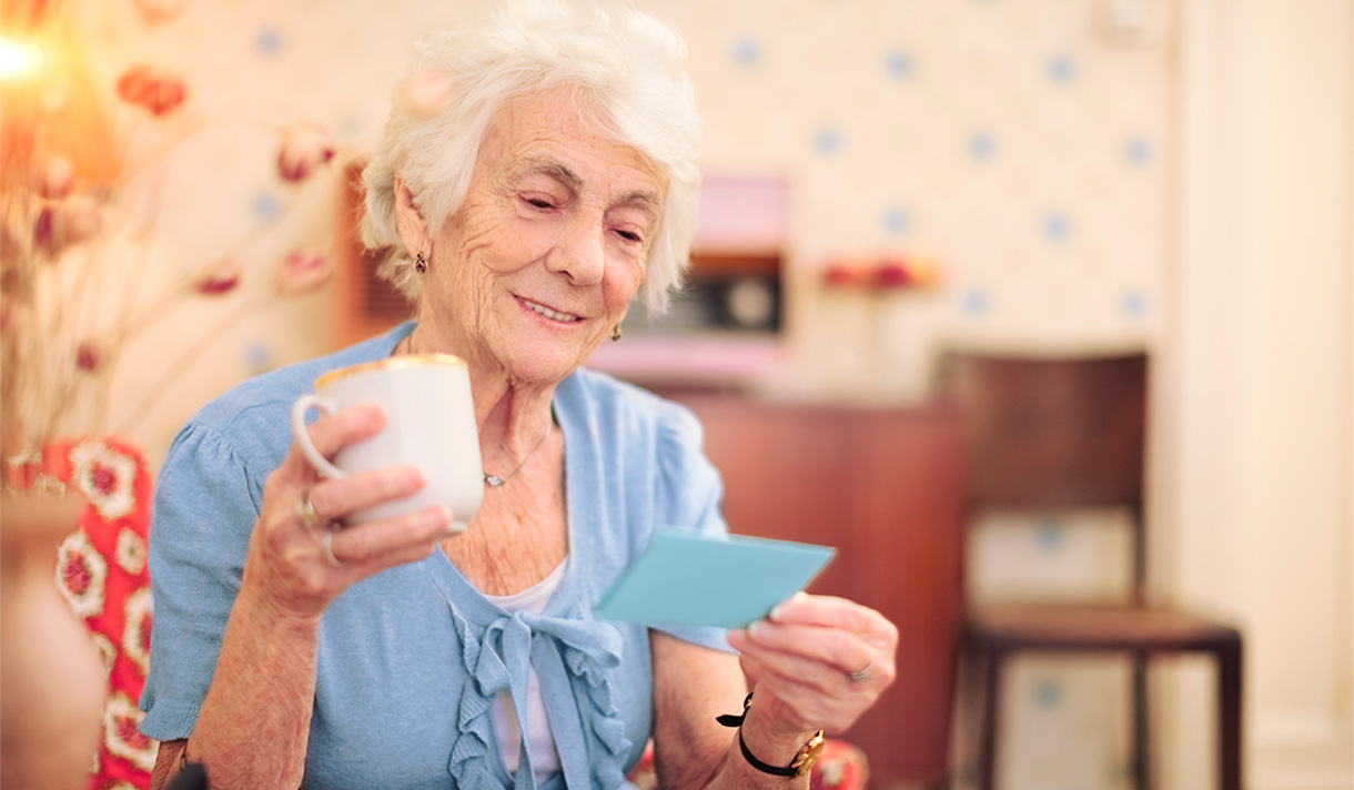 Elderly woman with holding coffee cup and smiling as she reads the card in her hand.