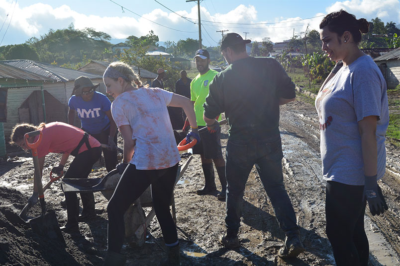 Students Katie Capobianco, Stefanie Campos, Melissa Mahadeo, Chanelle Sears, Robby O’Malley, and local Bridges to Community member, Victor, shovel gravel into wheelbarrows to create the cement.