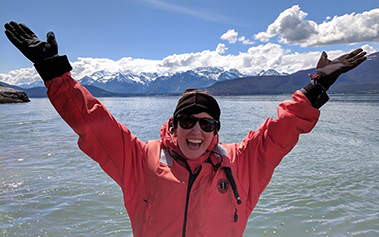 Alumni in parka standing in front of water and mountains with arms raised