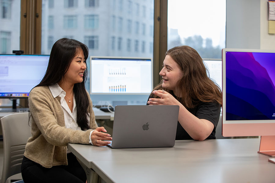 Two Ph.D. students discuss a project while sitting at a table  reviewing content on a laptop.