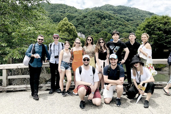 A photo of a group of study-abroad students posing for a photo on a bridge with a lush green mountain in the background.