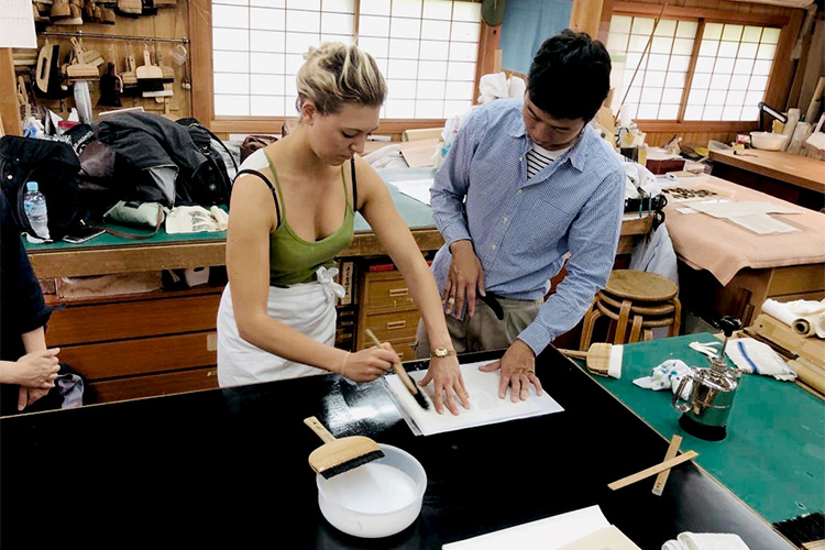 A photo of a traditional Japanese woodworking/paper-making workshop. Various tools and materials, including brushes and wooden frames, are laid out on a green table.