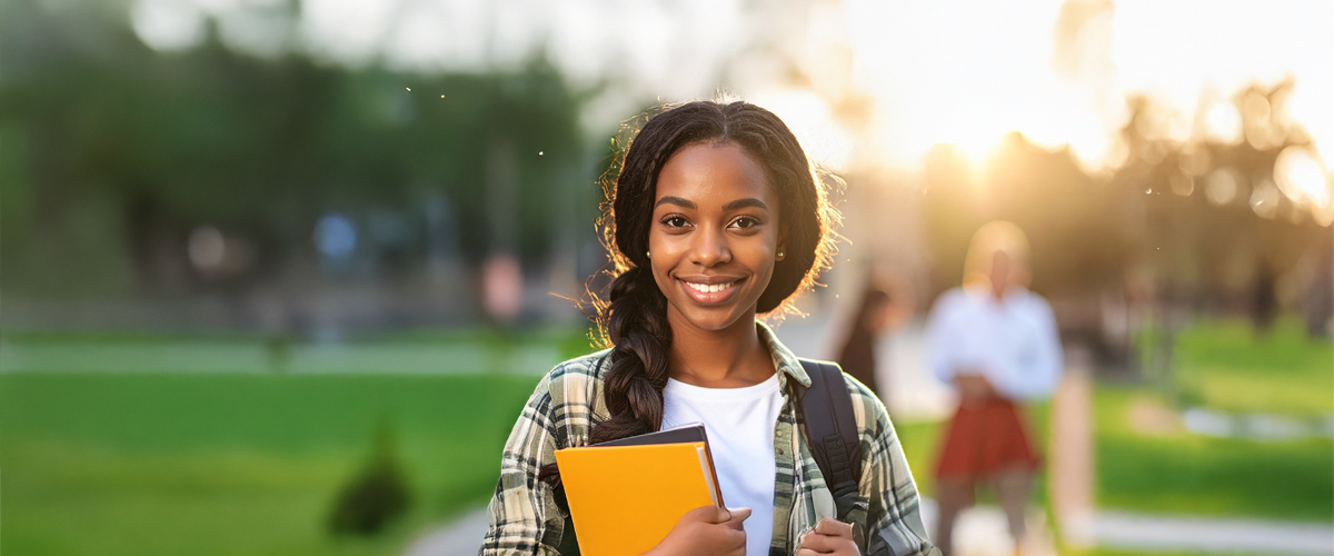 A student smiling. Holding a yellow notebook and has a backpack on in a quad