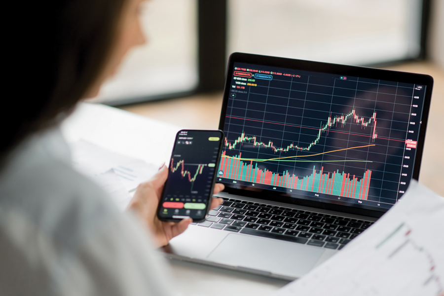 A woman on a laptop and phone viewing the same graph application and comparing it to a piece of paper