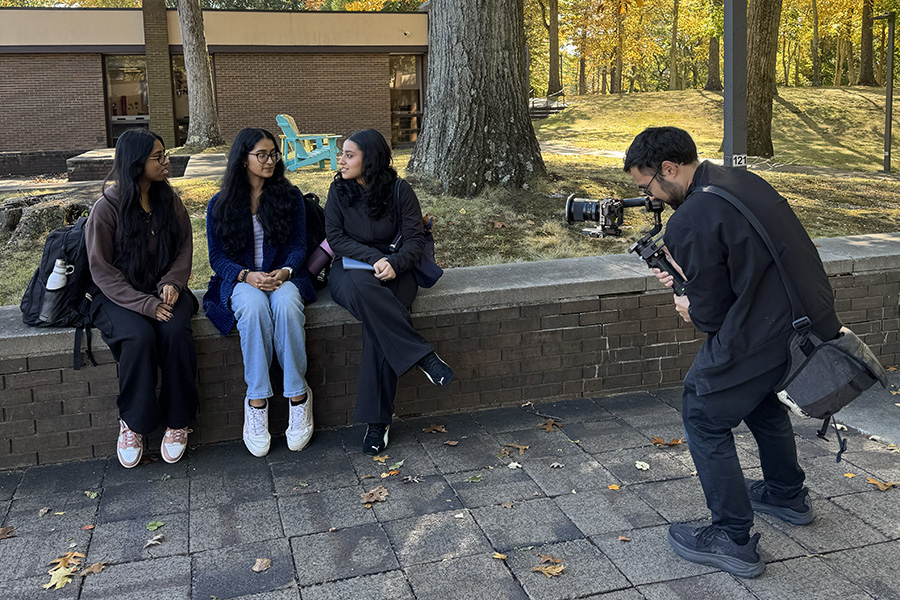 Three students sit on a bench while a videographer is filming there for a social media video.