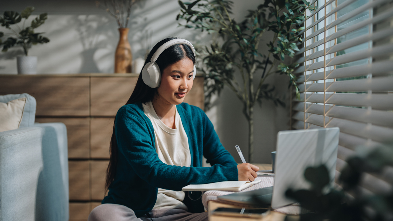 Woman wearing headphones sitting in front of a laptop and writing in a book