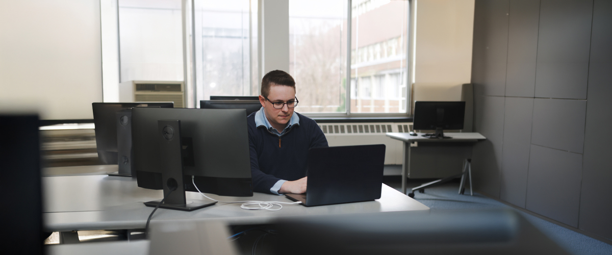 A graduate student works at a desk behind computer monitors and a laptop.
