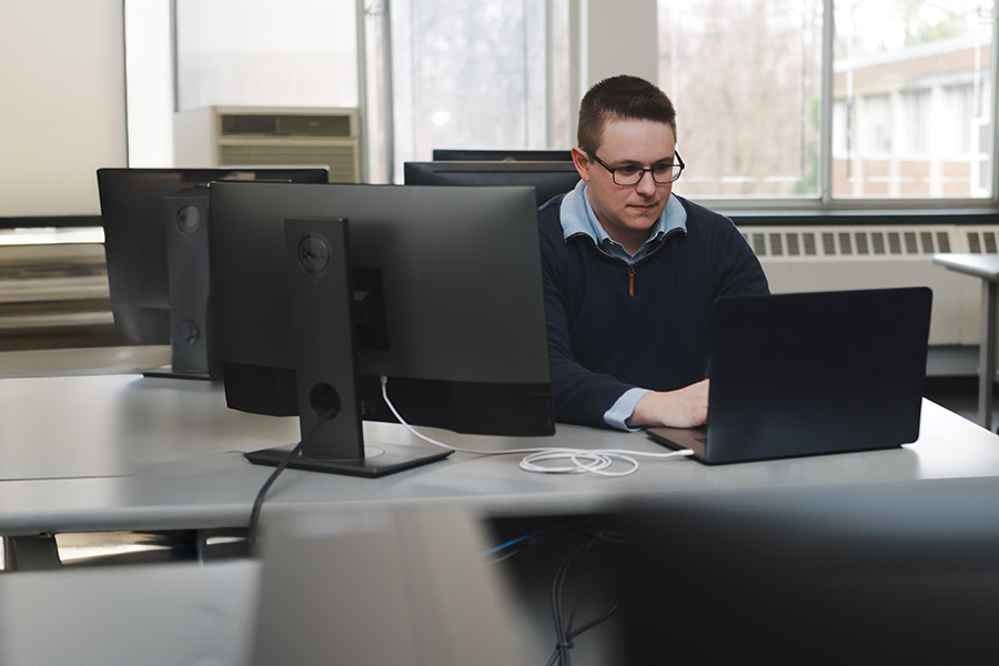 A graduate student works at a desk behind computer monitors and a laptop.