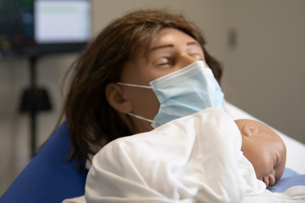 High-fidelity maternal manikin wearing a surgical mask and holding an infant manikin in a simulated hospital bed